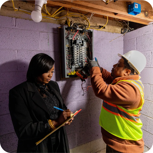 A woman writes on a clipboard while an electrician wearing a safety vest and hardhat works on a breaker box 